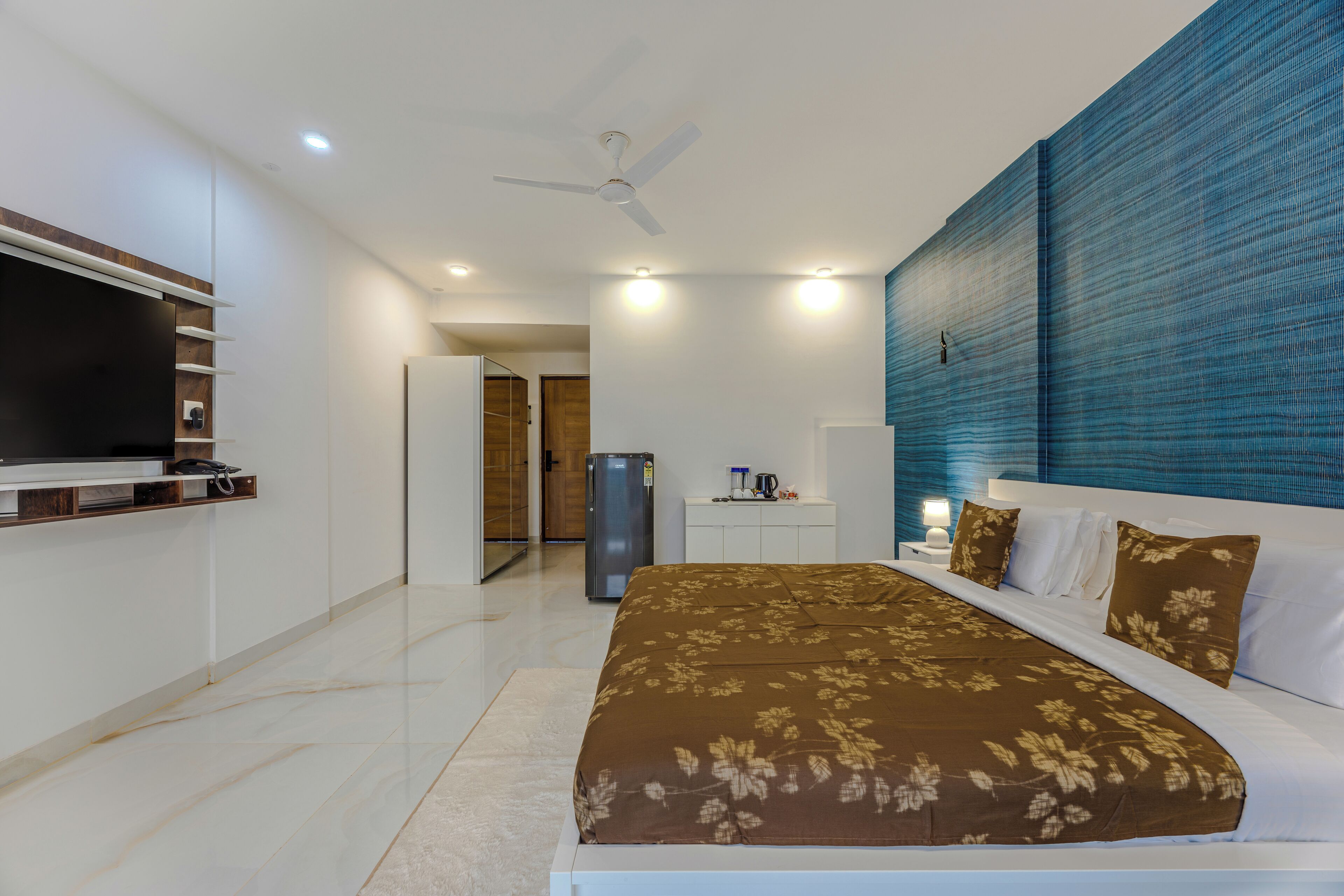 Quarto de hotel moderno com uma cama grande, televisão de tela plana, ventilador de teto e banheiro visível ao fundo.