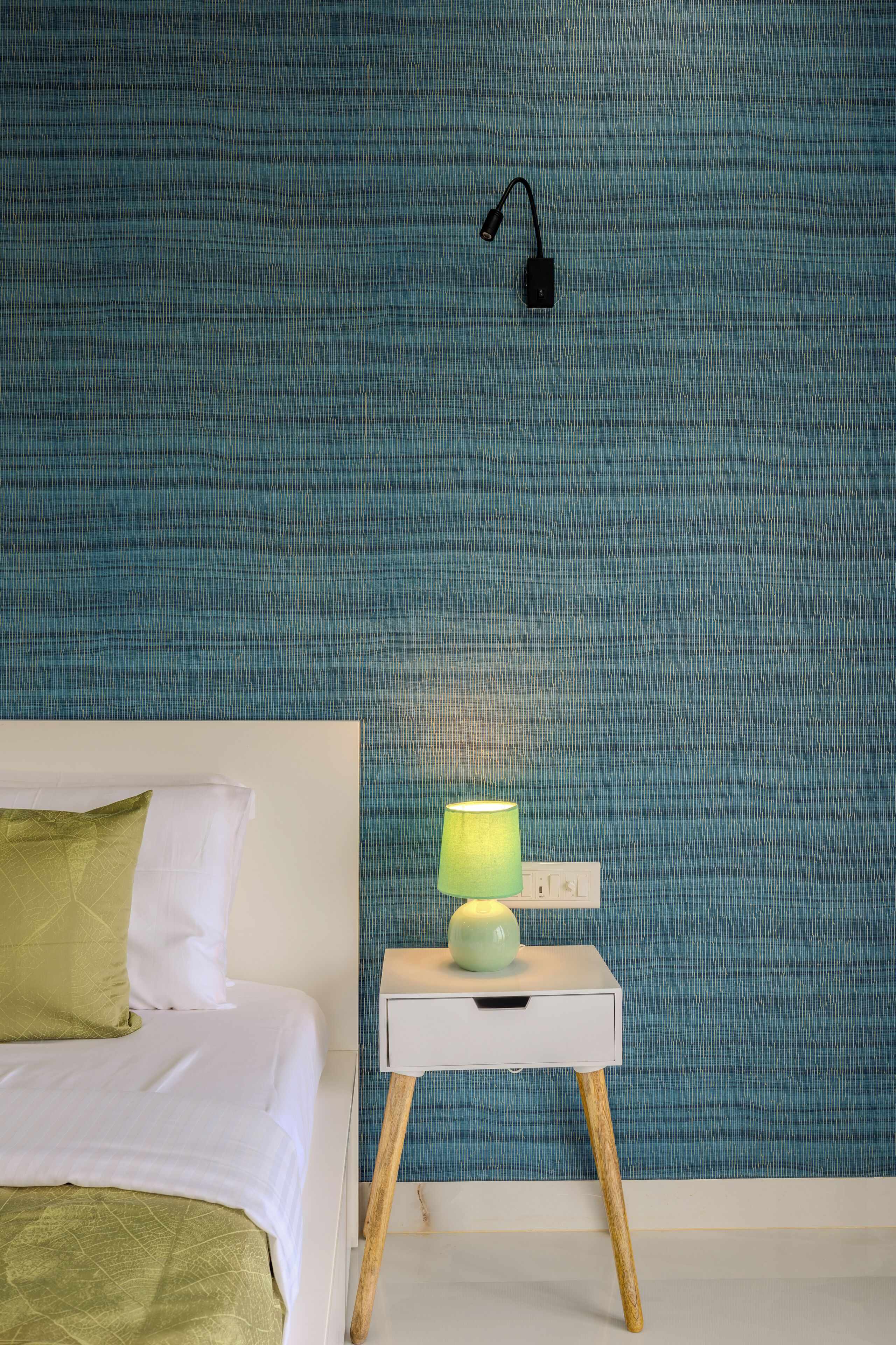 Una cama bien tendida con un almohadón verde y una manta blanca, una mesita de noche con una lámpara verde y una pared azul con textura.
