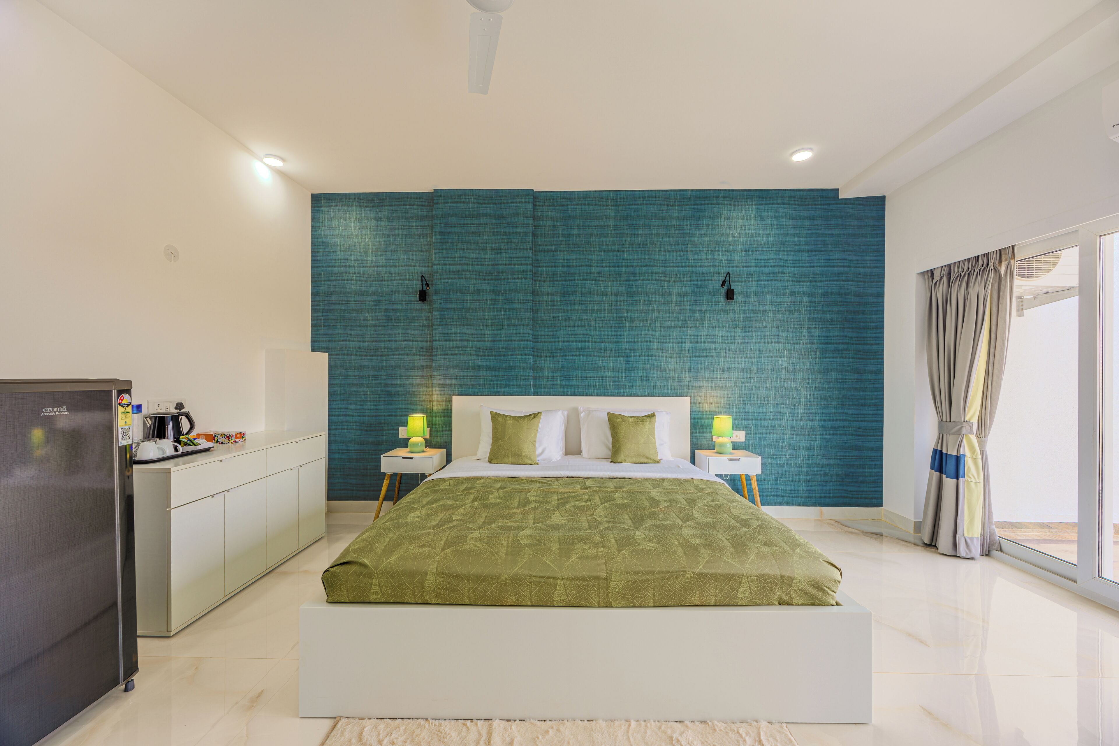 Une chambre moderne avec un grand lit, une armoire intégrée, un petit coin cuisine et une grande fenêtre avec des rideaux.