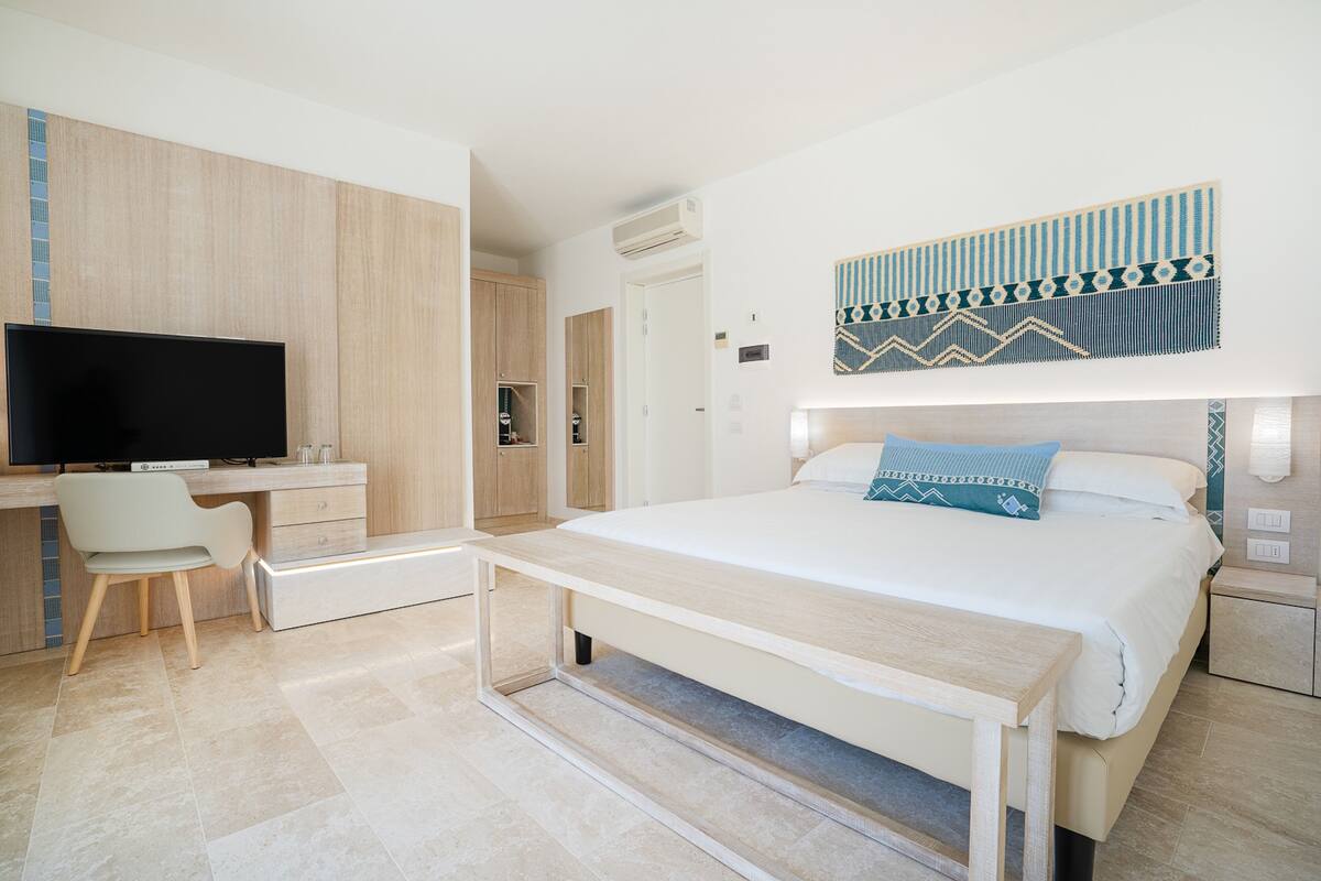 A modern hotel room with a bed, bedside table, TV, desk, chair, and a decorative wall hanging.