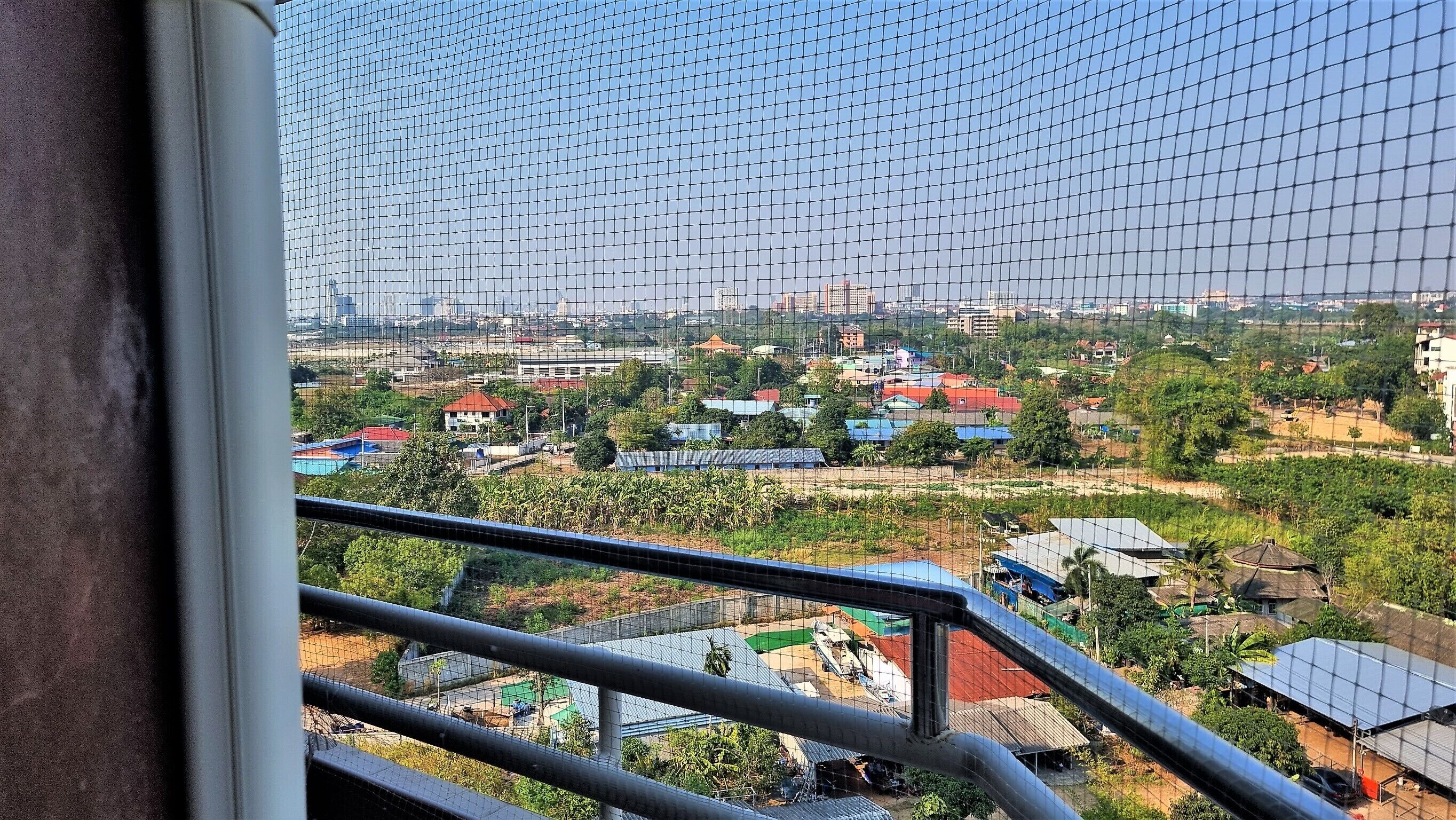 Blick durch ein Fenster mit einem Metallgitter auf ein Wohngebiet mit Häusern, Bäumen und einer entfernten Skyline.