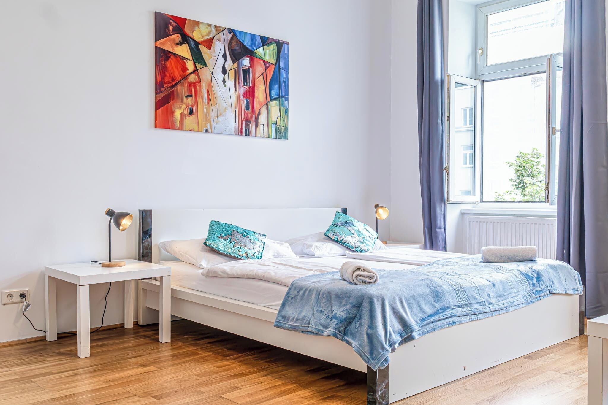 A modern bedroom with a white bed, two nightstands, a colorful abstract painting, and a window with curtains.