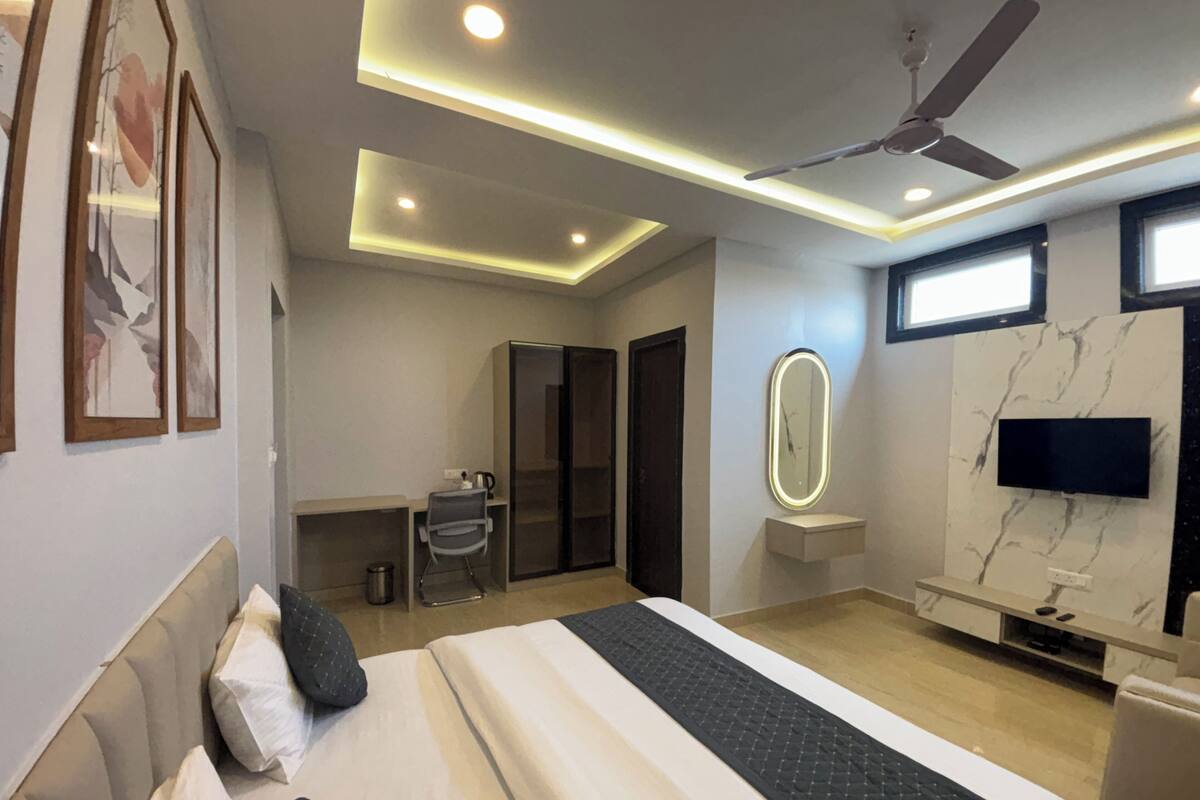 A modern hotel room with a large bed, a desk, a chair, a vanity with a mirror, a television, and a ceiling fan.