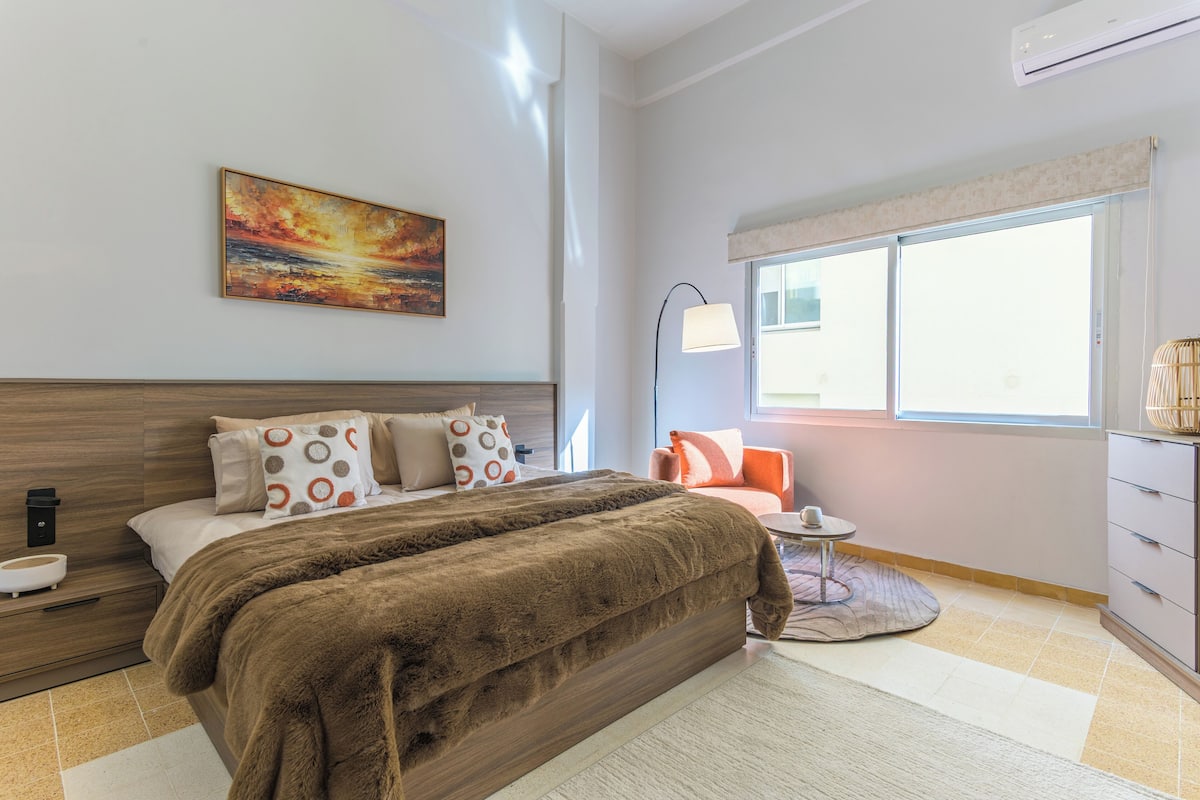 Ein modernes Schlafzimmer mit einem großen Bett, einem Nachttisch, einer Lampe, einem Sessel, einem kleinen Tisch und einem Fenster.