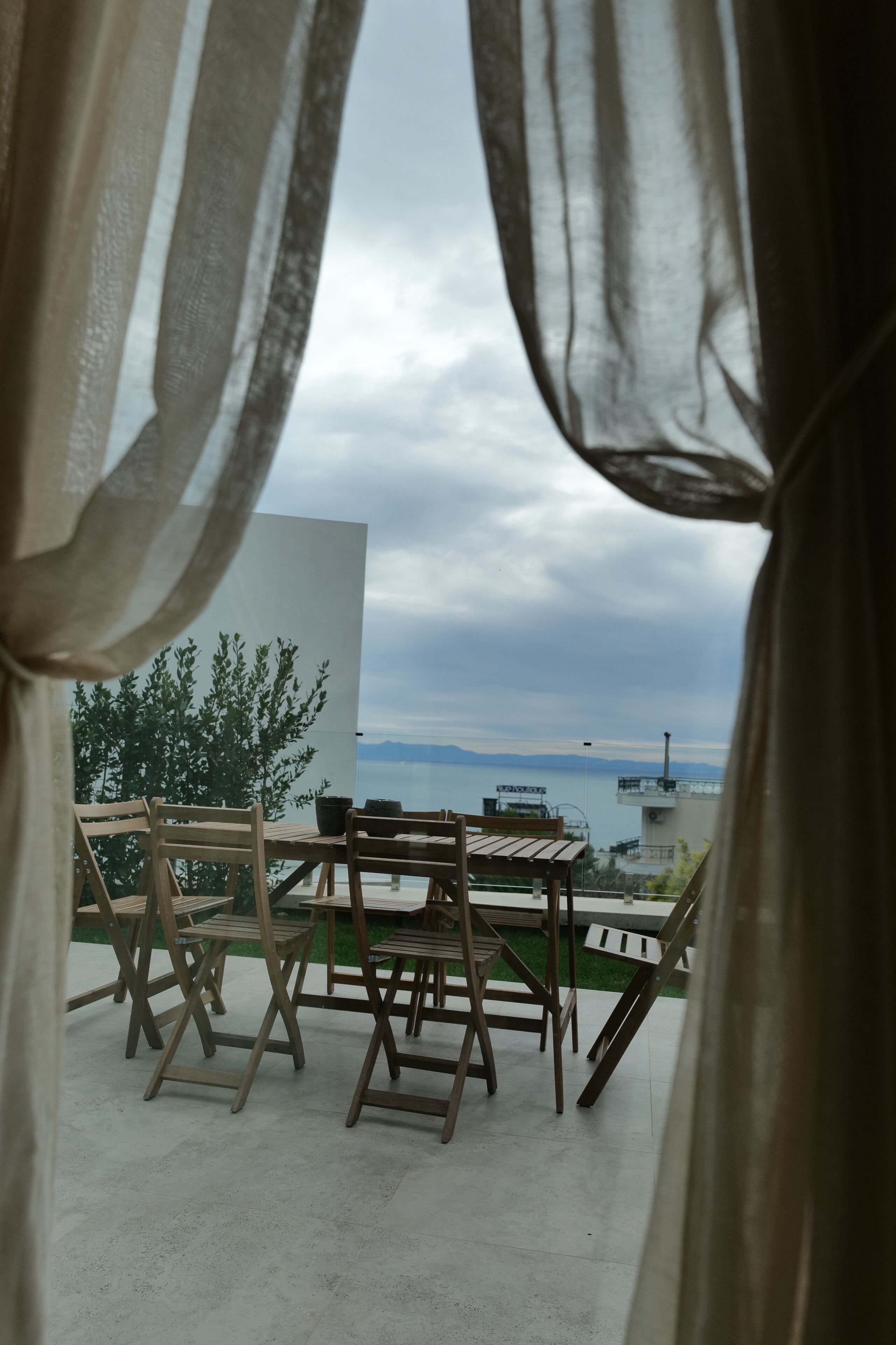 A balcony with wooden chairs and tables, overlooking a body of water and distant mountains.