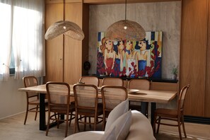 A dining area with a wooden table and chairs, a painting on the wall, and a hanging light fixture. - Stay By Ilvesta Dhermi (Dhërmi)