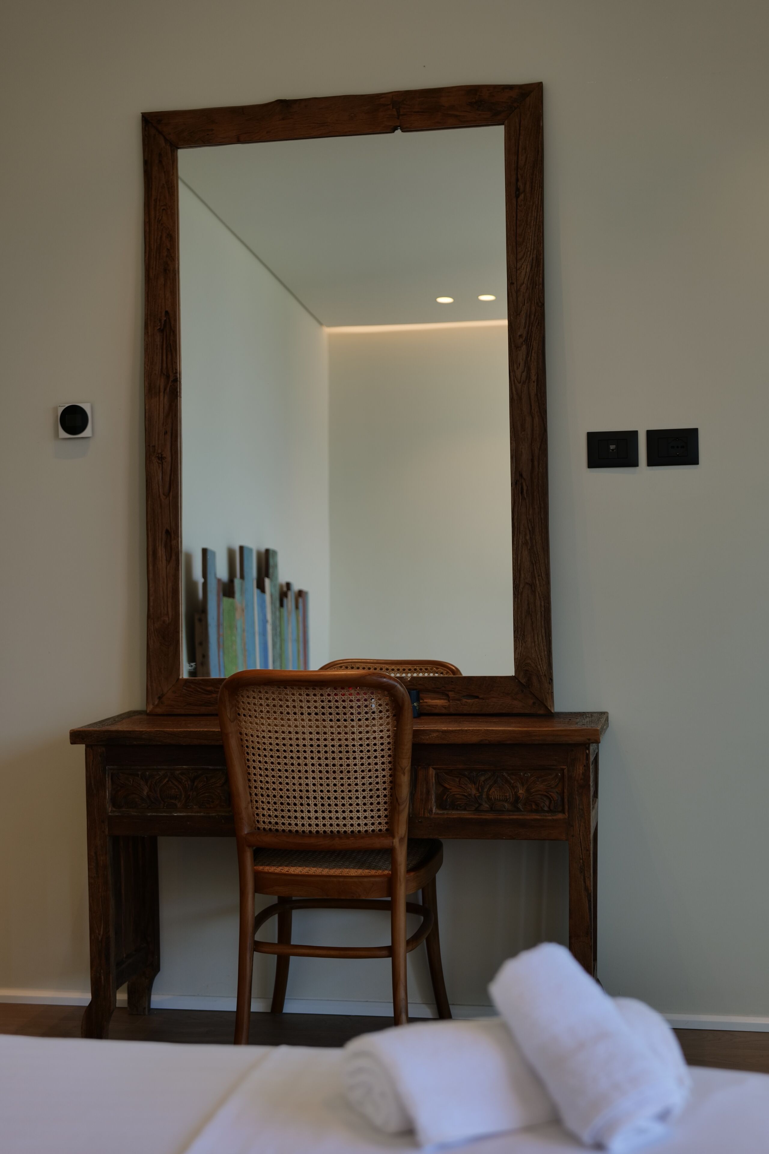 A wooden desk with a mirror, a wicker chair, and a neatly folded white towel on a bed.