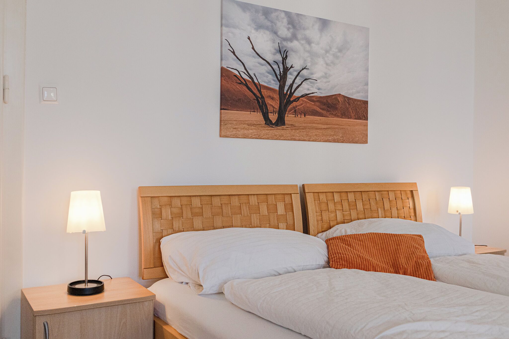 Une chambre d’hôtel avec un grand lit, deux tables de chevet, une lampe fixée au mur et un tableau représentant un paysage désertique.