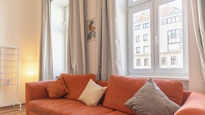 Ett modernt vardagsrum med en soffa, kuddar, en lampa och ett fönster med utsikt över byggnader.