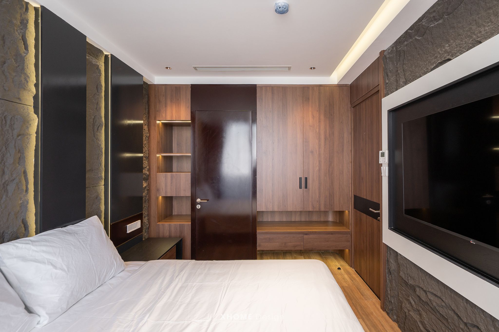 A modern bedroom with a large bed, built-in wooden shelving, a TV, and a stone accent wall.