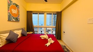 A hotel room with a bed, a ceiling fan, a window with curtains, and a view of buildings. - 1BR Service Apart Parel 2503 Near TMH By Florastays (Mumbai)