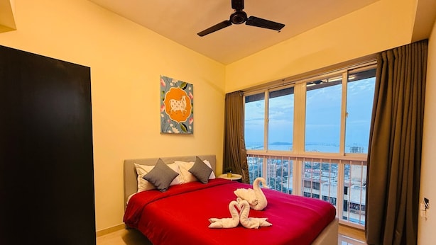 A bedroom with a bed, a ceiling fan, a window with a view of the city, and a painting on the wall. - 1BR Service Apart Parel 2503 Near TMH By Florastays (Mumbai)