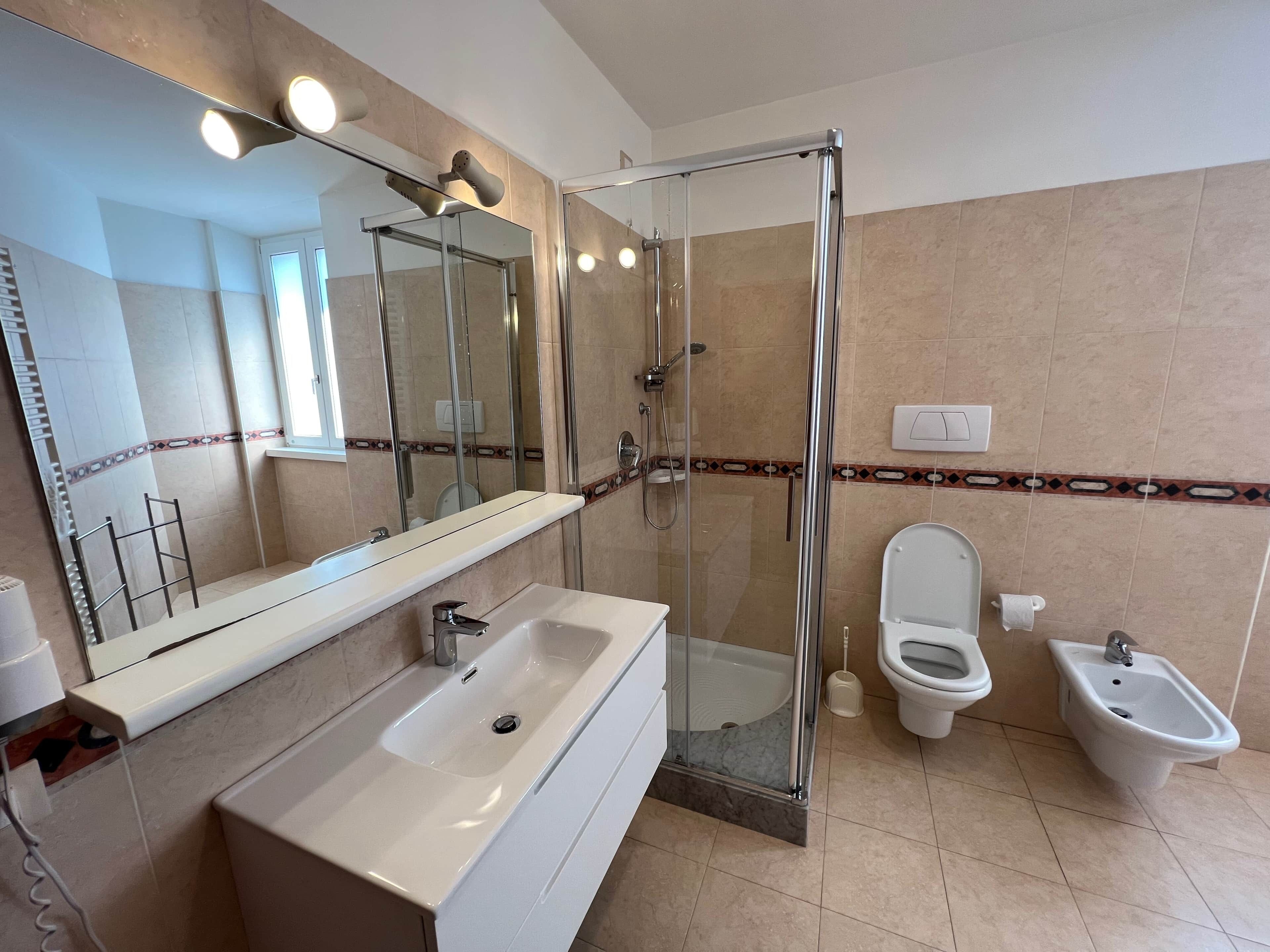 Een moderne badkamer met een douche met glazen wand, een grote spiegel, een wastafel, toilet en een bidet.