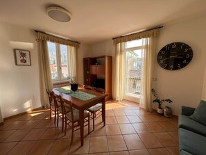 A dining area with a wooden table and chairs, a clock on the wall, and a view through the window. - Superior 3 (Nago-Torbole)
