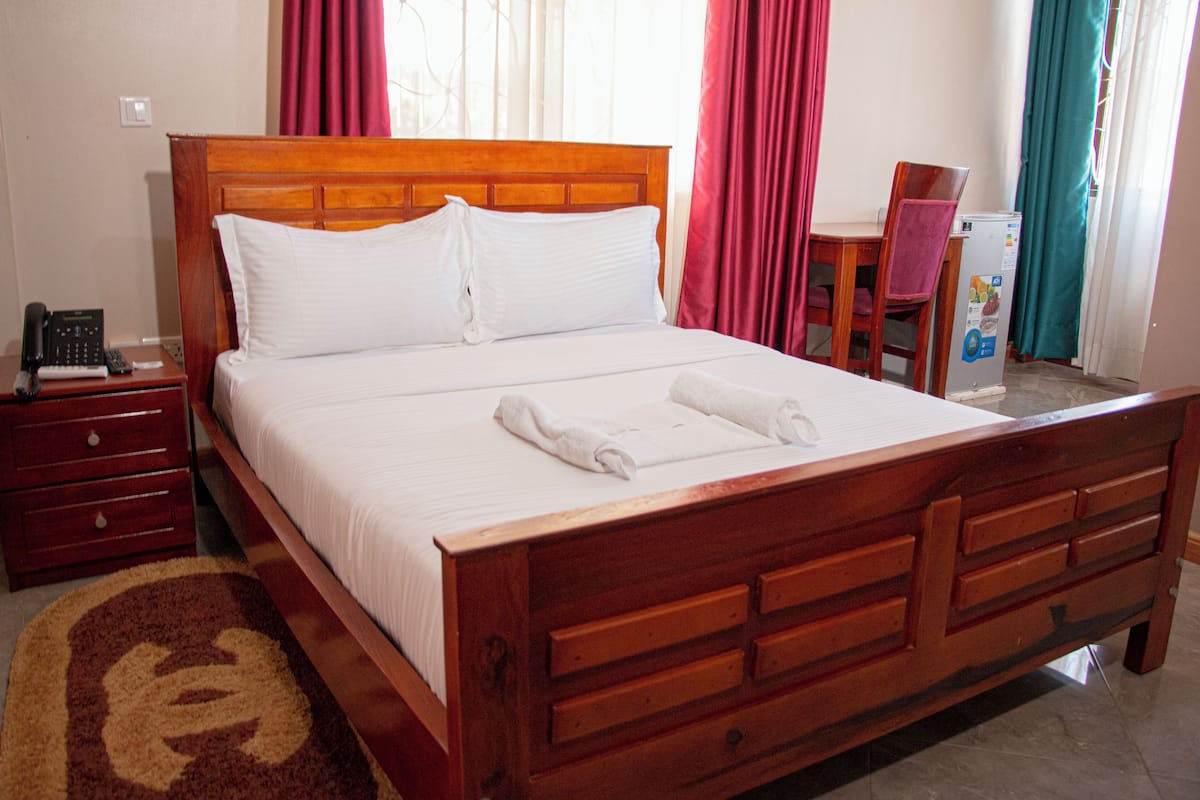A hotel room with a wooden bed, a nightstand with a phone, a chair, and a small table with a refrigerator.
