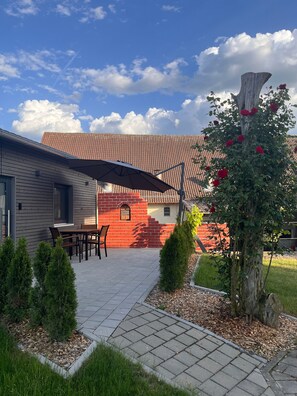 Ein Gebäude mit einem Ziegeldach, einer Terrasse mit Tisch und Stühlen sowie einem Garten mit Baum und Sträuchern.