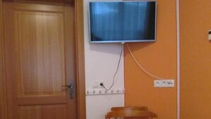 Une petite pièce avec une porte en bois, un téléviseur fixé au mur, une chaise en bois et deux lits avec du linge de lit jaune.