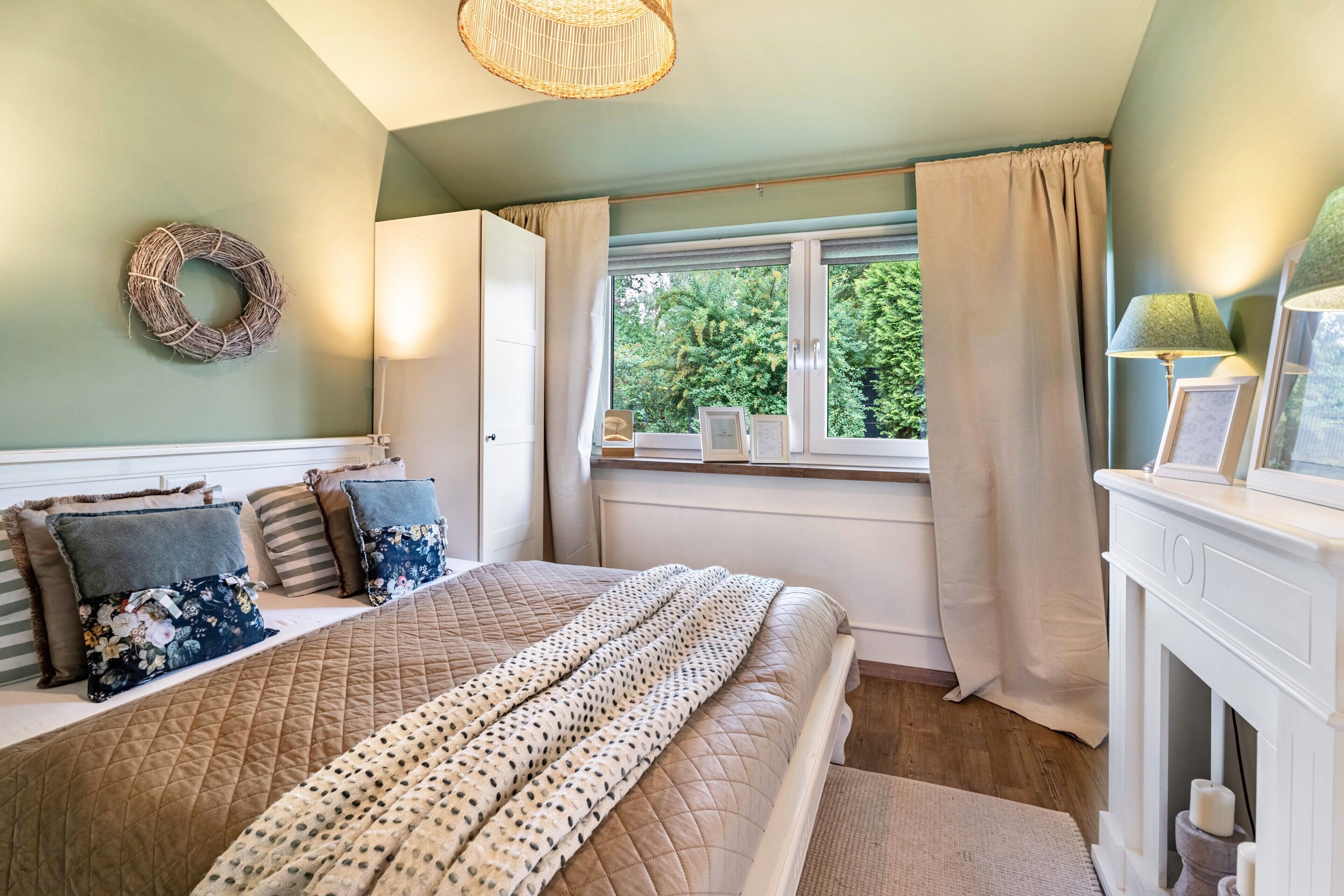 A bedroom with a bed, a window with curtains, a fireplace, and a wall-mounted wreath.