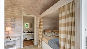 Une chambre avec un plafond en bois, un lit avec des rideaux à rayures, une table de chevet avec une lampe, une fenêtre donnant sur un beau paysage et une étagère intégrée où sont rangées des serviettes.