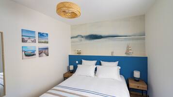 Une chambre avec un grand lit, une tête de lit ornée d’un panneau bleu et un tableau représentant un paysage marin. On y trouve quatre tableaux encadrés accrochés au mur, deux tables de chevet avec des lampes et un luminaire suspendu.