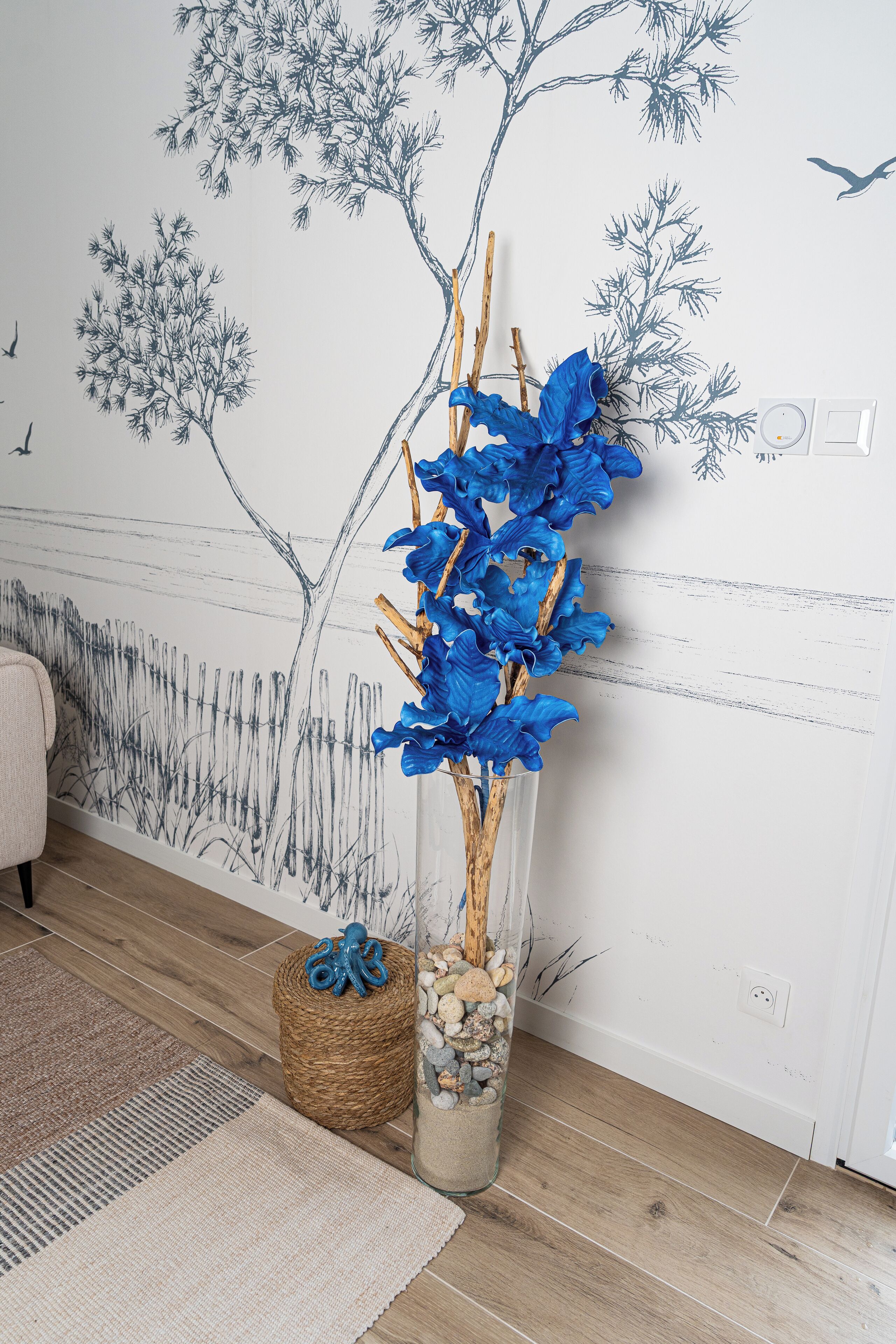 Une pièce dont un mur est décoré d’une fresque représentant des arbres et des oiseaux, avec un vase contenant des fleurs bleues, un panier en osier et une petite statue.