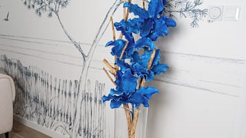 Une pièce dont un mur est décoré d’une fresque représentant des arbres et des oiseaux, avec un vase contenant des fleurs bleues, un panier en osier et une petite statue.