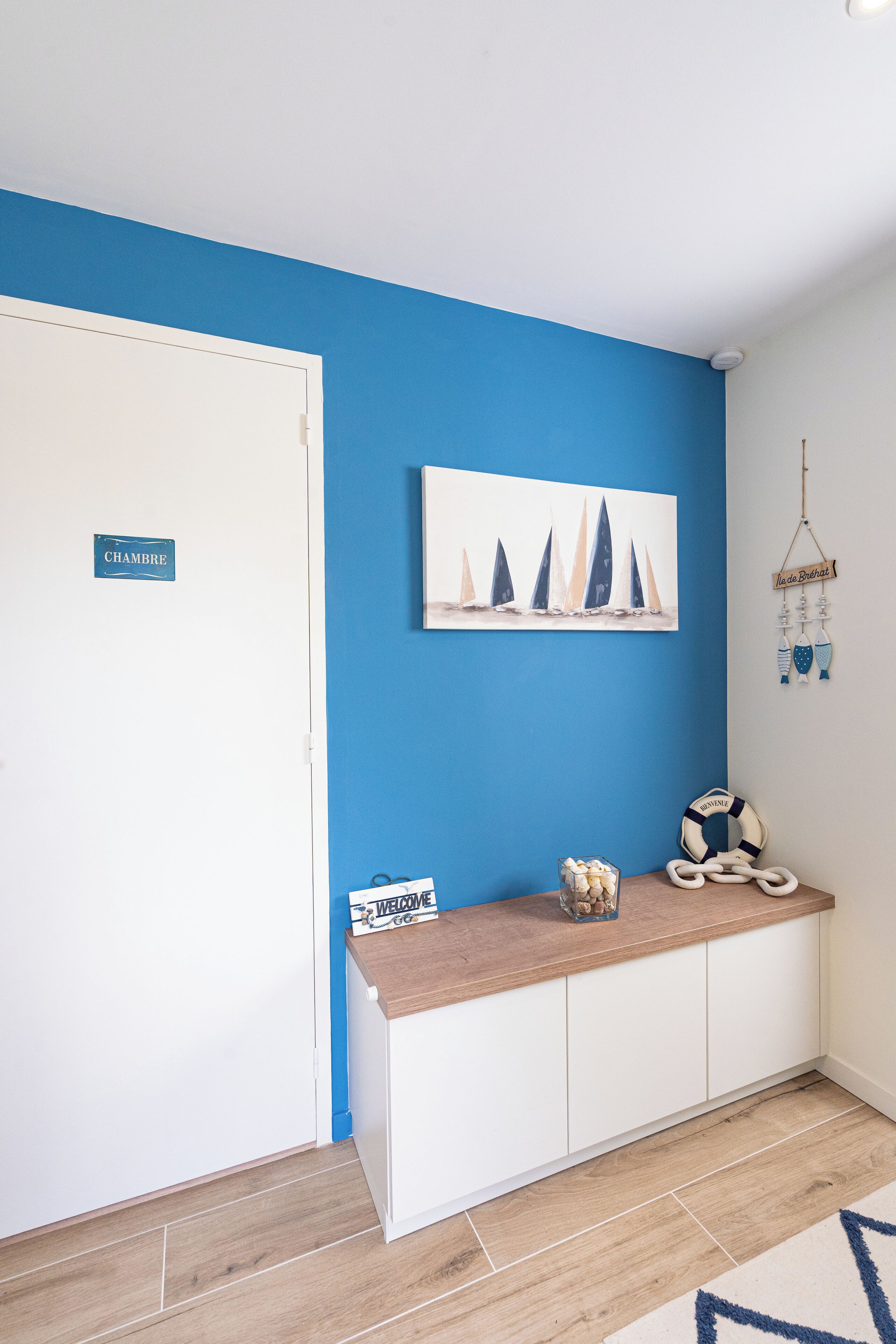 Un área de recepción con una pared azul, una puerta blanca con la leyenda 'CHAMBRE', un cartel de bienvenida, un adorno de salvavidas y una pintura con temática náutica.