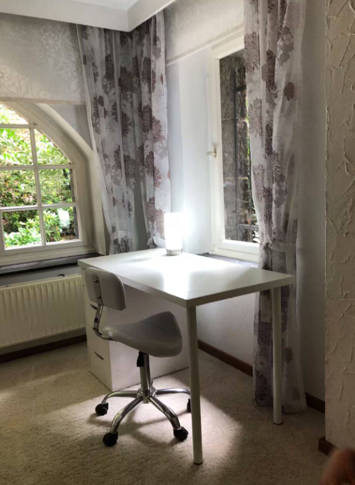 Ein Zimmer mit weißem Schreibtisch, Stuhl, Fenster mit Vorhängen und Heizkörper.