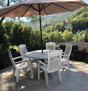 Un patio con una mesa redonda y cuatro sillas, una sombrilla y vistas a una ladera boscosa.