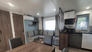 A compact living space with a dining area, a sofa, a kitchen with a microwave, and a window with curtains. - Bungalow between nature and ocean at Le Ferret, a 4* resort with aquatic areas (Lège-Cap-Ferret)