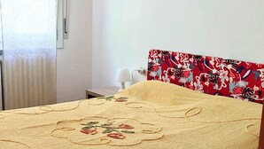 Een bed met een bloemenprint sprei, een rood gestreept hoofdbord, een bedlamp en een raam met gordijnen.