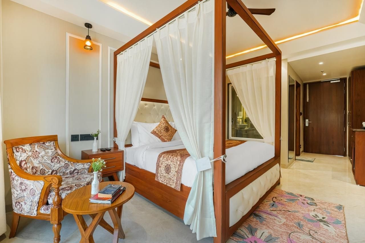 A four-poster bed with a canopy, a wooden chair, a small table with books, and a bedside lamp.