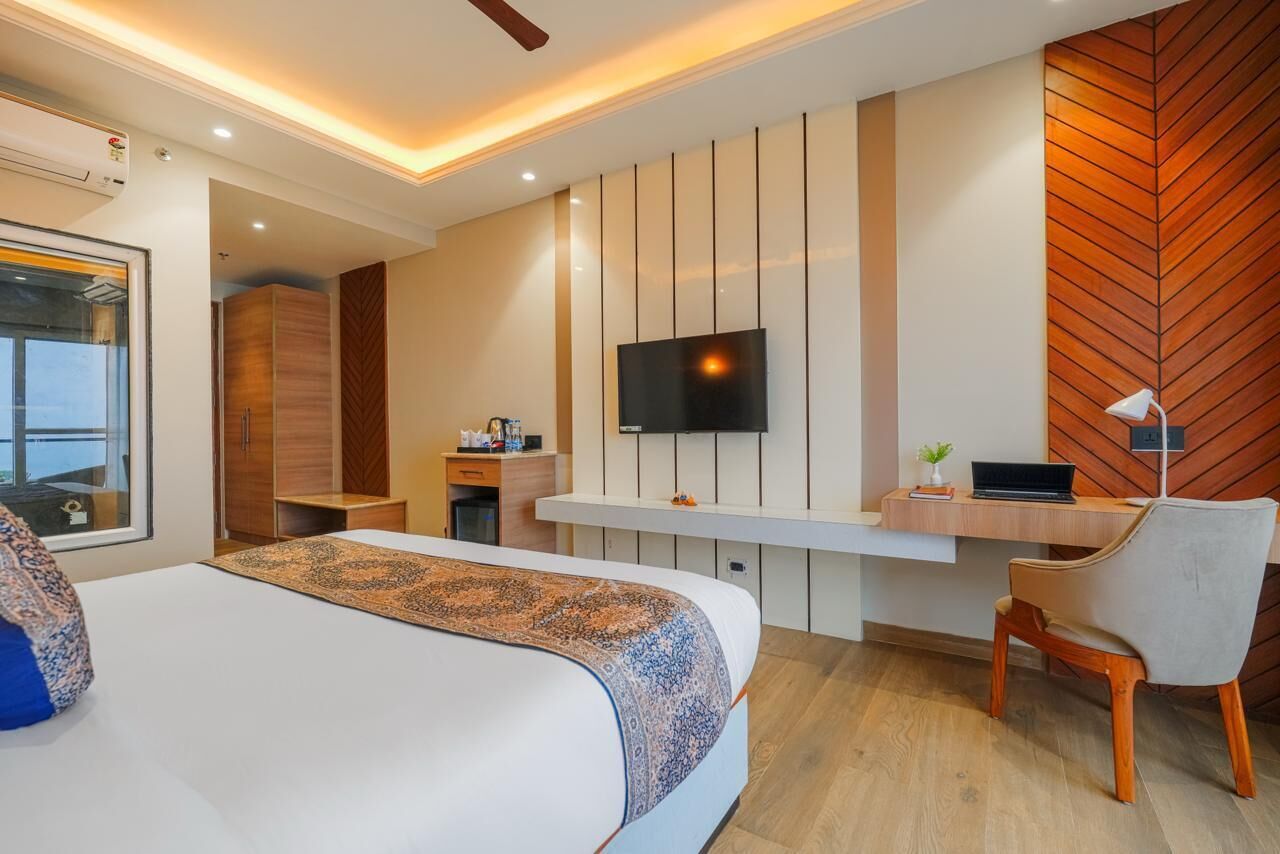 A modern hotel room with a large bed, a desk with a laptop, a TV mounted on the wall, and a herringbone-patterned wall.