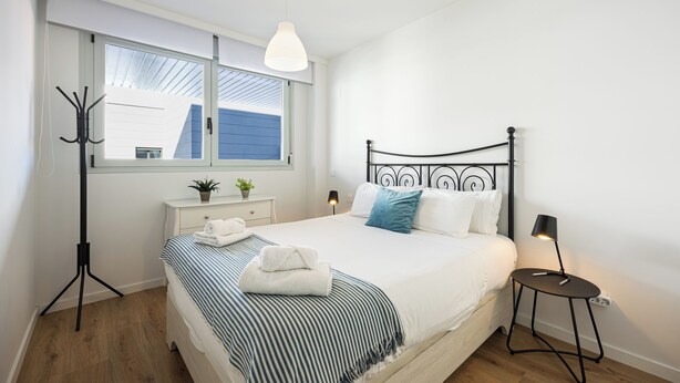 A bedroom with a bed, bedside table, lamp, and a window with a view of a blue building. - RivasCenter Apartment (Rivas-Vaciamadrid)