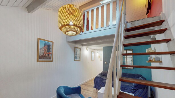 Une pièce avec un lit en mezzanine, une chaise bleue, un escalier en bois et un luminaire suspendu.