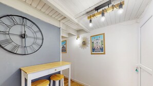Une pièce avec une grande horloge murale, une table en bois et deux tabourets, ainsi que des tableaux encadrés accrochés au mur.