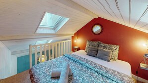 Une chambre avec un puits de lumière, un lit avec une literie à motifs, un mur d’accent rouge et une petite table de chevet avec une lampe.