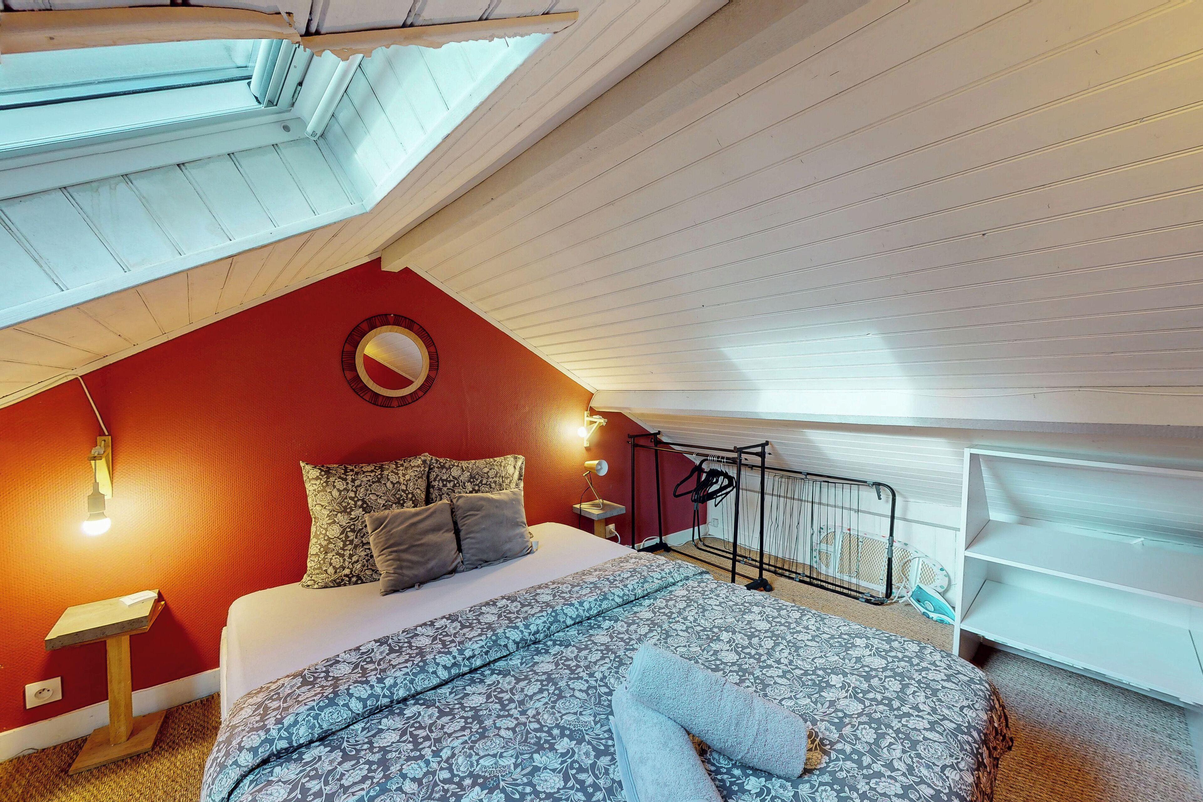 A bedroom with a sloped ceiling, a bed with patterned bedding, a round mirror, and a small table with a lamp.
