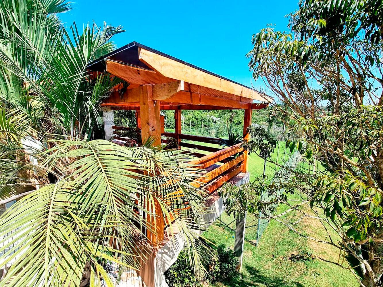 A wooden gazebo with a sloped roof, surrounded by lush greenery and palm trees.