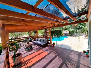 Eine Holzterrasse mit einer Couch und Topfpflanzen, mit Blick auf einen Swimmingpool.