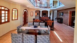 A spacious living area with a glass coffee table, patterned sofas, and a high ceiling with a red staircase.