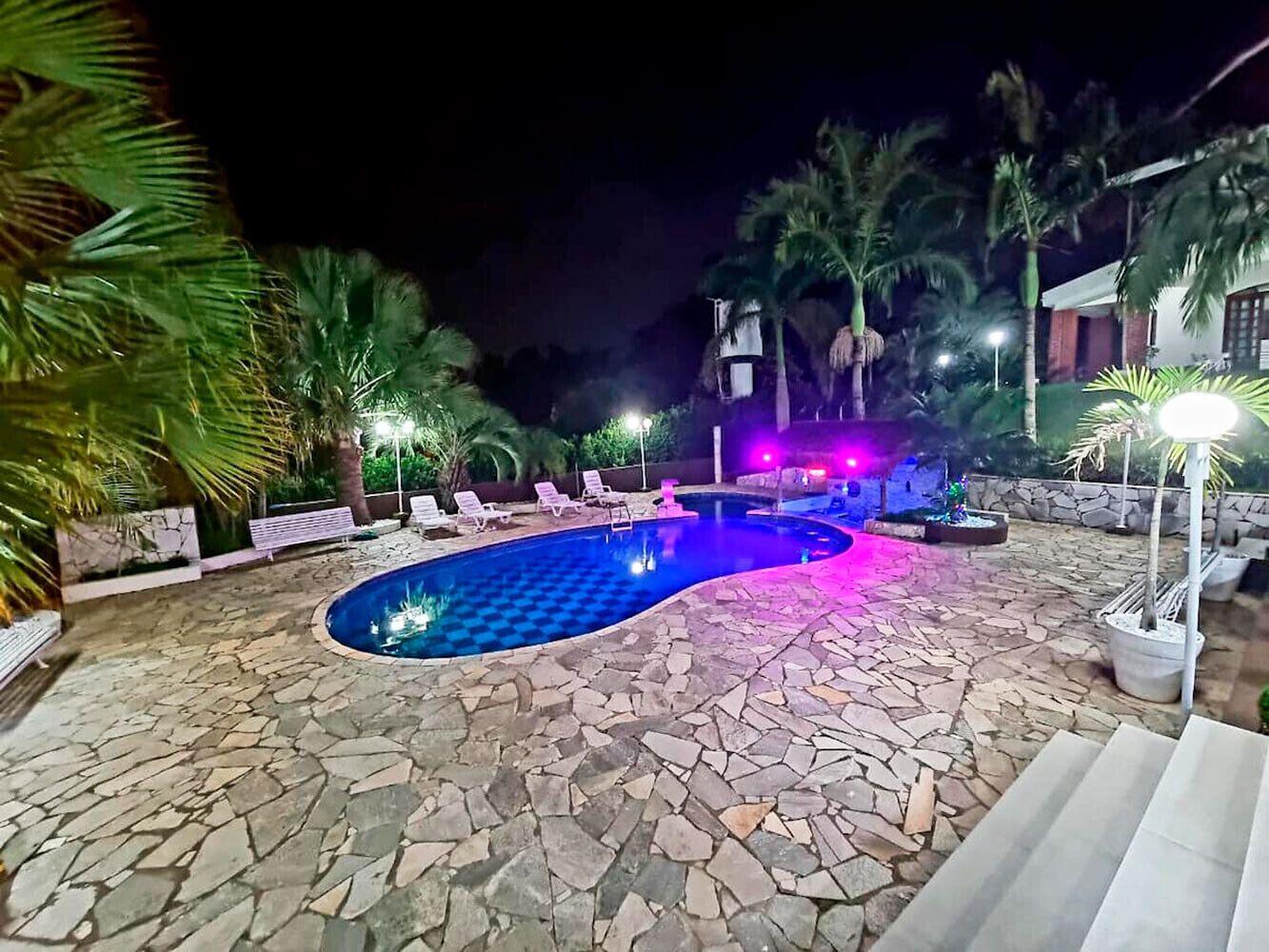 Zona de piscina iluminada de noche con suelo de piedra, sillas de descanso y palmeras.