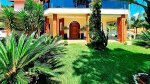 A modern two-story house with a red door, surrounded by lush greenery and palm trees.