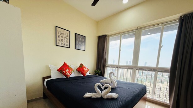 A hotel room with a large bed, two red floral pillows, a dark blue bedspread, and a view of a cityscape through large windows. - 1BR Service Apart Parel 3405 Near TMH By Florastays (Mumbai)