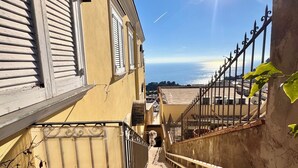 Una escalera conduce a un balcón con vista al mar, barandilla y un edificio con contraventanas.