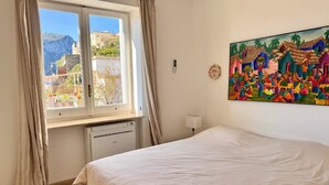 Un dormitorio con cama, ventana con cortinas, radiador, lámpara de pared y un cuadro de colores.