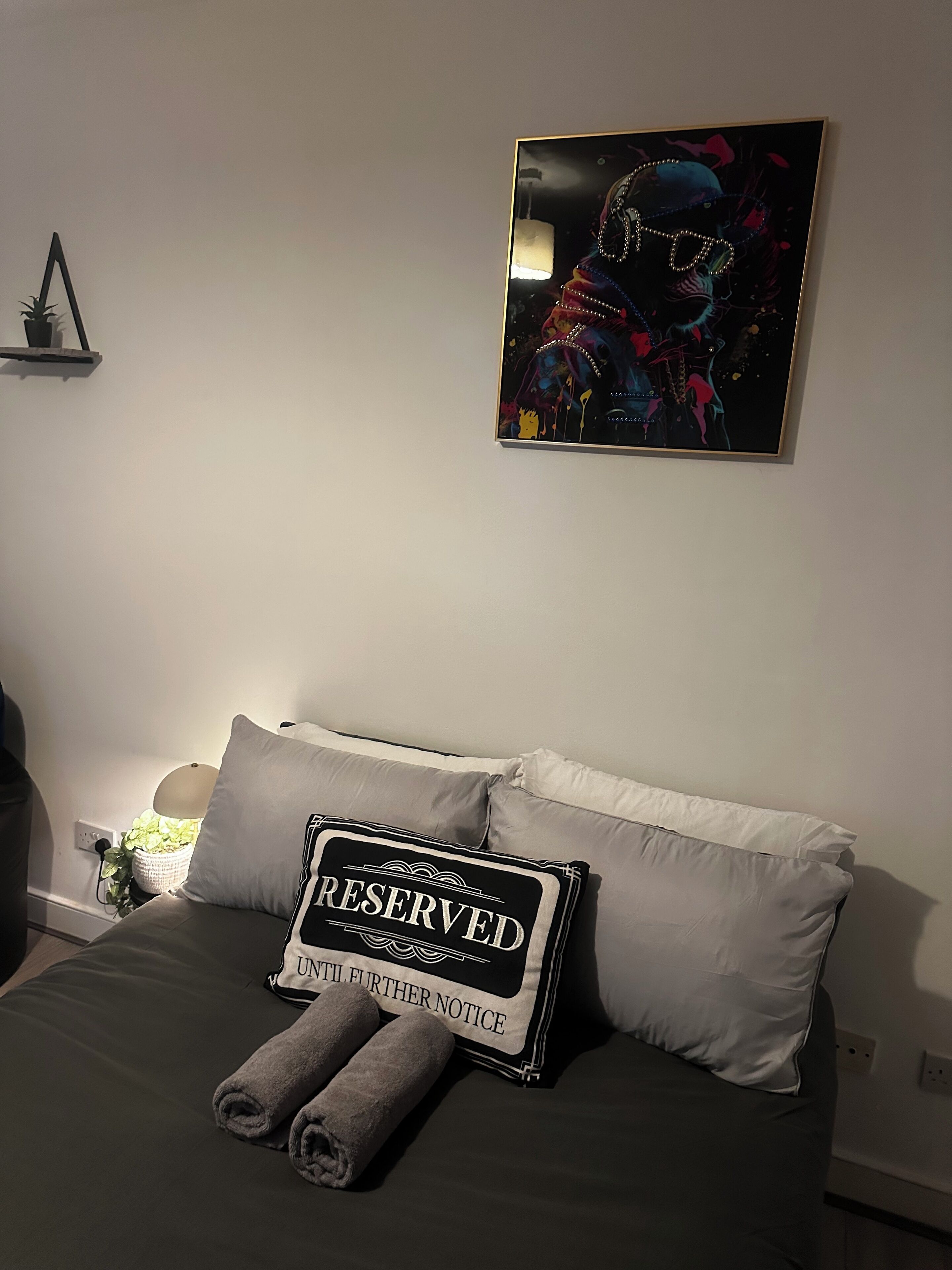 A bedroom with a bed, pillows, a framed artwork, and a bedside table with a lamp.