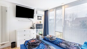 Quarto moderno com televisão, cómoda branca, duas camas com roupa de cama estampada e uma janela grande com persianas.