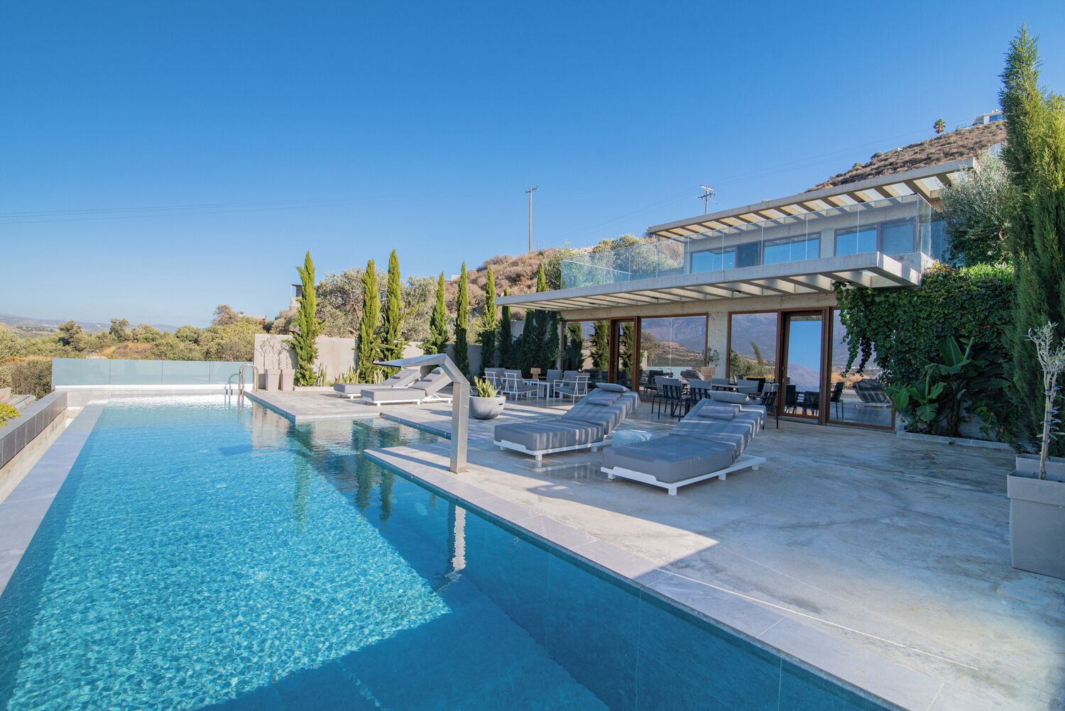 Un área de piscina moderna al aire libre con sillones de descanso y una zona de estar cubierta, rodeada de vegetación y con vistas a un paisaje pintoresco.