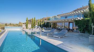 Un área de piscina moderna al aire libre con sillones de descanso y una zona de estar cubierta, rodeada de vegetación y con vistas a un paisaje pintoresco.